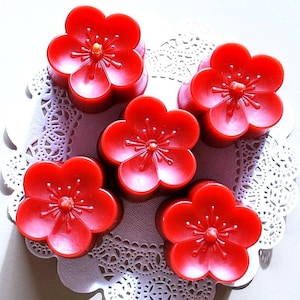 May include: Five red wax candles shaped like cherry blossoms. The candles are arranged on a white doily.