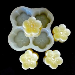 May include: A silicone mold for making five small, flower-shaped candles. The mold is white and has a textured surface. The candles are a pale yellow color and are in the shape of a five-petal flower.
