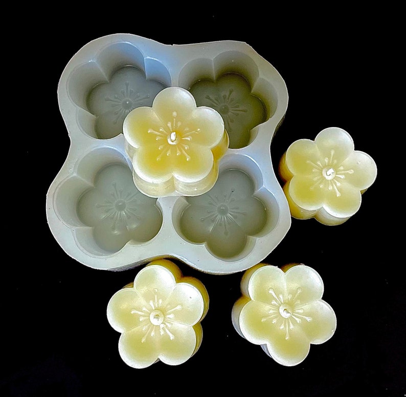 May include: A silicone mold for making five small, flower-shaped candles. The mold is white and has a textured surface. The candles are a pale yellow color and are in the shape of a five-petal flower.
