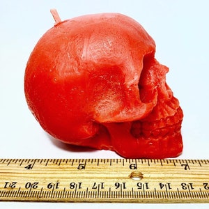 May include: A red skull-shaped candle with a wick on top. The candle is about 6 inches long and is sitting on a wooden ruler.