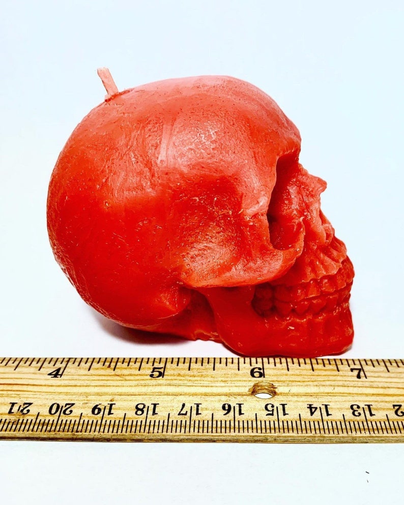 May include: A red skull-shaped candle with a wick on top. The candle is about 6 inches long and is sitting on a wooden ruler.