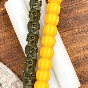 Puede incluir: Un conjunto de cuatro velas cónicas. Dos son lisas y blancas, una es de color verde oliva con un diseño de calaveras apiladas y una es de color amarillo brillante con una textura segmentada similar a una calabaza. Cada vela tiene una mecha.