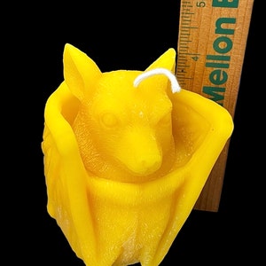 May include: A yellow beeswax candle shaped like a bat. The bat is curled up with its wings wrapped around its body. The candle has a white wick.