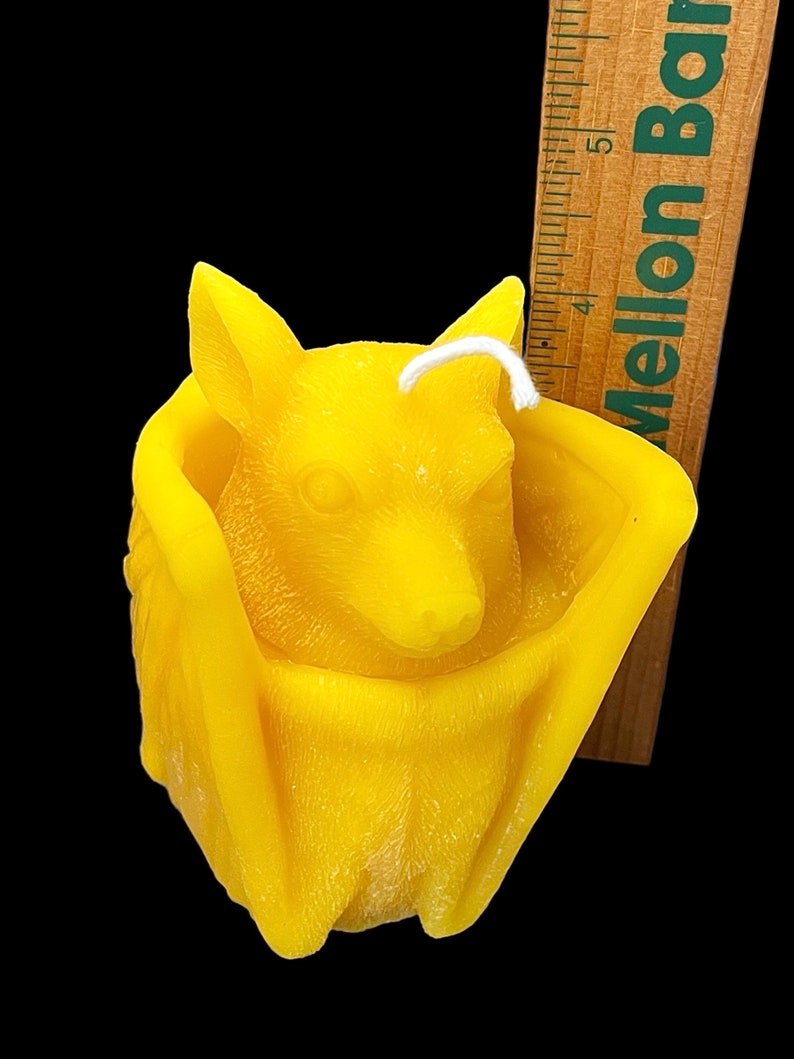 May include: A yellow beeswax candle shaped like a bat. The bat is curled up with its wings wrapped around its body. The candle has a white wick.