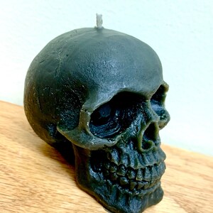 May include: A dark green skull-shaped candle with a white wick. The candle is detailed with a realistic skull design, including eye sockets, a nose, and teeth.