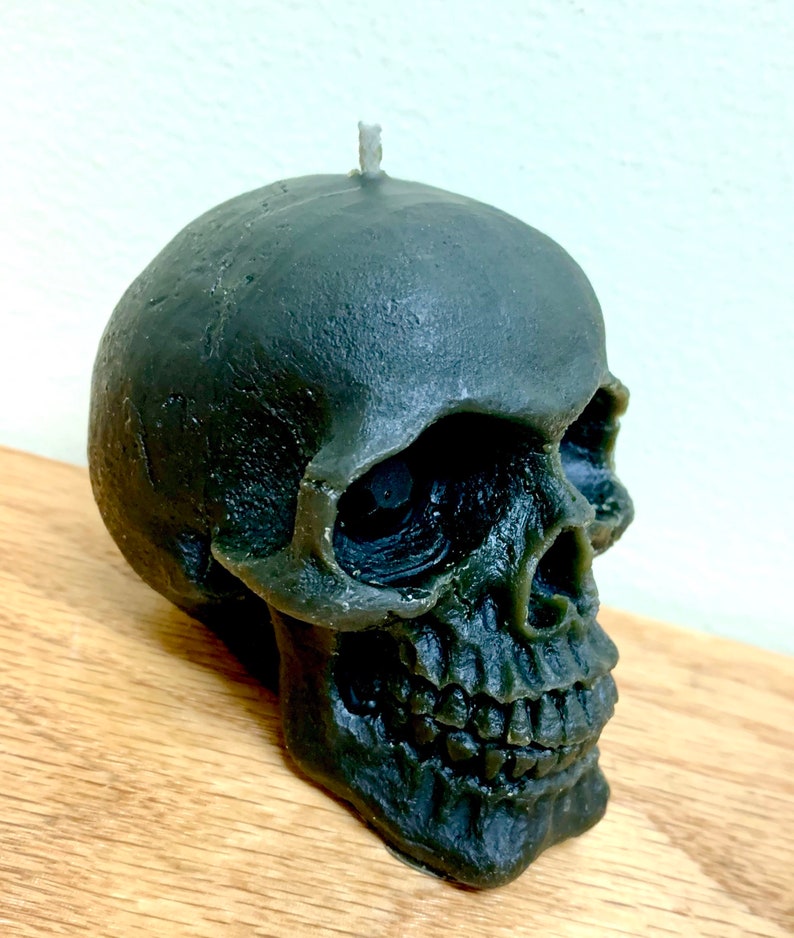 May include: A dark green skull-shaped candle with a white wick. The candle is detailed with a realistic skull design, including eye sockets, a nose, and teeth.