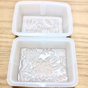 May include: Two white silicone soap molds. The top mold has a design of bees and honeycomb. The bottom mold has a design of sunflowers and leaves.