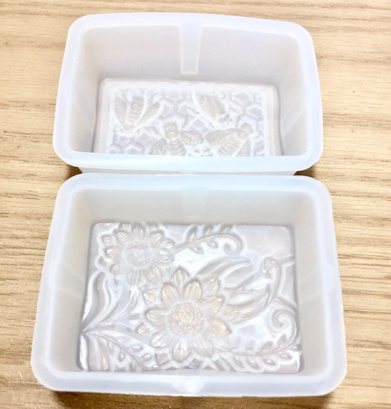 May include: Two white silicone soap molds. The top mold has a design of bees and honeycomb. The bottom mold has a design of sunflowers and leaves.
