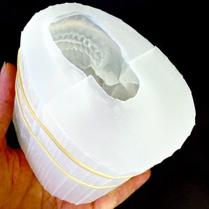 May include: A white silicone mold for making resin crafts. The mold is shaped like a skull and has a detailed impression of teeth. The mold is held in a hand and is secured with rubber bands.