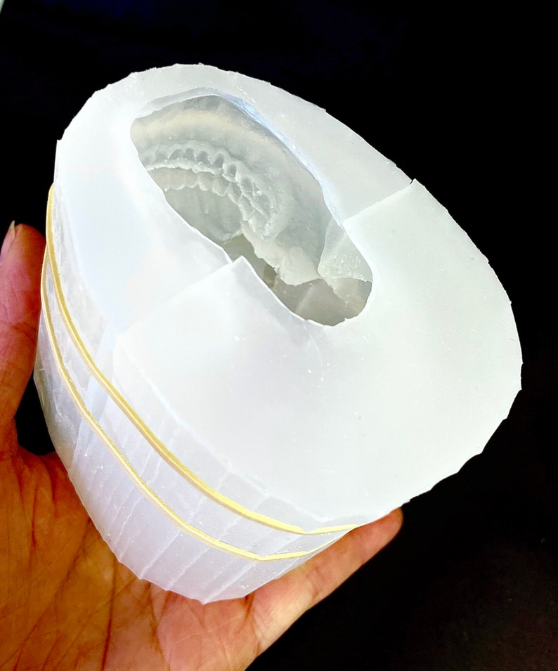 May include: A white silicone mold for making resin crafts. The mold is shaped like a skull and has a detailed impression of teeth. The mold is held in a hand and is secured with rubber bands.