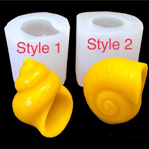 以下が含まれることがあります： 貝殻の形をしたキャンドルを作るための白いシリコンモールド2つ。モールドには「スタイル1」と「スタイル2」のラベルが貼られています。モールドの隣には、黄色い貝殻の形をしたキャンドルが2つ置かれています。