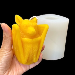 May include: A yellow wax candle shaped like a bat, with wings folded around its body. The candle is being held in a hand, and a white silicone mould is visible in the background.