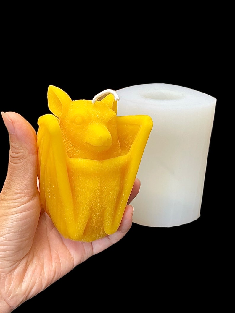 May include: A yellow wax candle shaped like a bat, with wings folded around its body. The candle is being held in a hand, and a white silicone mould is visible in the background.