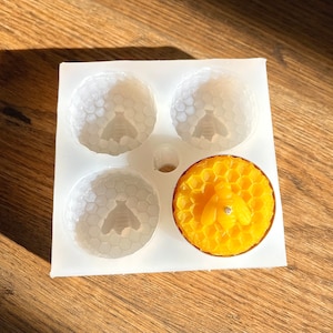 May include: A white silicone mold with four round cavities, each with a honeycomb pattern. A yellow beeswax candle with a honeycomb pattern and a bee design is in one of the cavities.