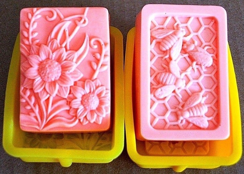 May include: Two pink soap bars in yellow silicone molds. One bar features a floral design with sunflowers and vines. The other bar features a honeycomb design with bees.
