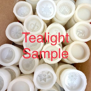 Puede incluir: Una colección de moldes de vela de té blanco con varios diseños, incluyendo un copo de nieve, una flor de lis, una colmena y un pájaro. Los moldes están dispuestos en una caja de cartón con el texto "Tealight Sample" impreso en la parte delantera.