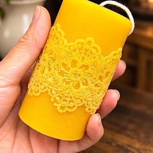 May include: A cylindrical yellow beeswax candle featuring a raised, intricate lace-like floral pattern wrapped around the center. The candle has a smooth, matte finish and a single white cotton wick protruding from the top edge.