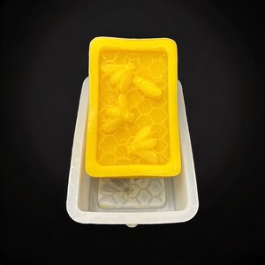 May include: A white silicone mold for making soap, with a yellow soap bar inside. The soap bar has a honeycomb pattern and bees on it.