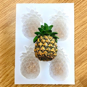 Könnte beinhalten: Eine weiße Silikonform mit vier Ananasformen. Eine goldene Ananasfigur wird in die Mitte der Form gelegt.