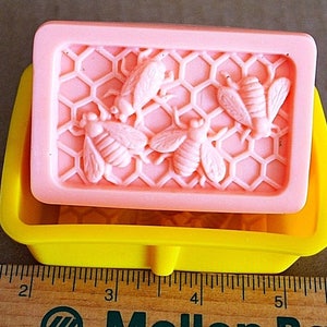 May include: A pink silicone soap mold with a honeycomb design and five bees. The mold is sitting on a yellow silicone base.