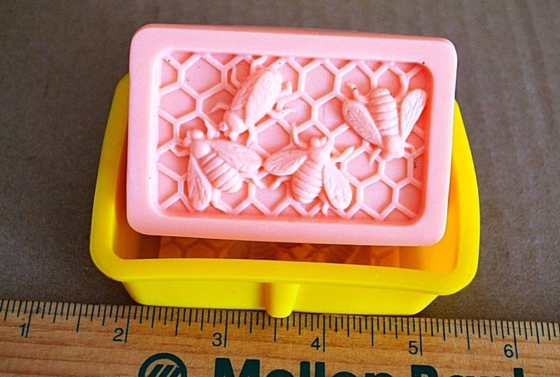 May include: A pink silicone soap mold with a honeycomb design and five bees. The mold is sitting on a yellow silicone base.