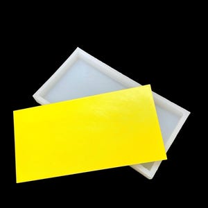 Pode incluir: Uma forma de silicone branca para fazer barras de sabão retangulares. A forma está aberta e uma barra de sabão amarela brilhante está colocada dentro.