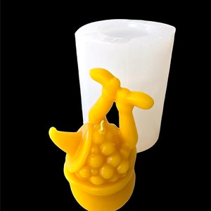 May include: A yellow candle shaped like a person sitting in a bucket with their legs up in the air. The candle is in a clear silicone mould.