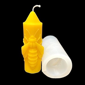 May include: A yellow beeswax candle with a bee design and a white wick. The candle is next to a clear silicone mold.