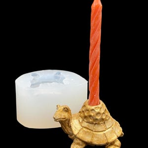 May include: A gold-coloured turtle candle holder with an orange spiral candle. A white silicone mould is next to the candle holder. The candle holder is a decorative item.