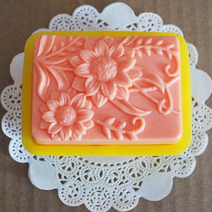 May include: A rectangular pink soap bar with a floral design featuring sunflowers and leaves. The soap is in a yellow silicone mold.