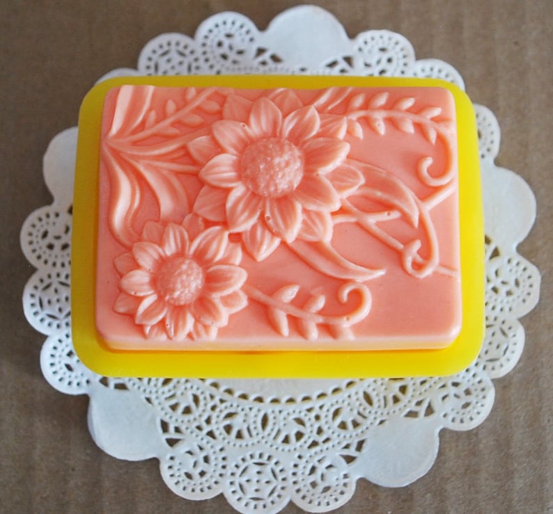 May include: A rectangular pink soap bar with a floral design featuring sunflowers and leaves. The soap is in a yellow silicone mold.