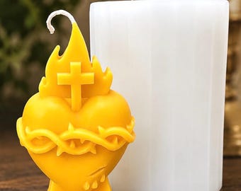 May include: A yellow candle shaped like a heart wrapped in a crown of thorns with a cross and flames on top, positioned next to a white cylindrical silicone mold with vertical ridges. The candle features a white wick extending from the flame.
