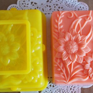 May include: Two handmade soap bars with floral designs. One bar is yellow with a raised sunflower design and the other is peach with a raised sunflower design.