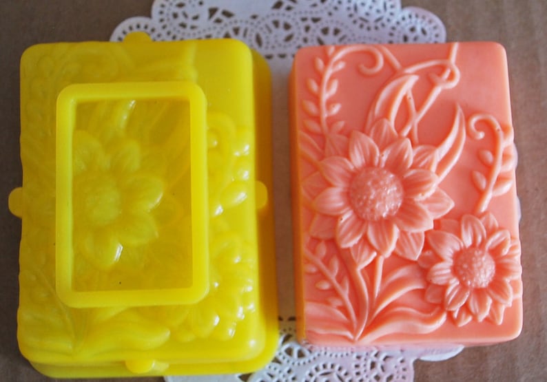 May include: Two handmade soap bars with floral designs. One bar is yellow with a raised sunflower design and the other is peach with a raised sunflower design.