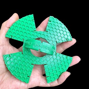May include: A green, 3D-printed object with a honeycomb pattern, held in a hand against a black background. The object is composed of four fan-shaped pieces and a central ring. The text "Handcrafted Clay" is visible on the ring.