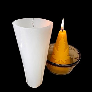 Könnte beinhalten: Eine weiße Silikon-Kerzenform in Form eines Kegels. Eine brennende gelbe Kerze mit einem grünen Blattdesign steht in einer durchsichtigen Glasschale.