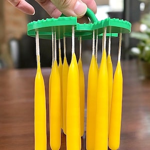 May include: A green plastic candle holder with a handle, holding eleven yellow candles. The candles are suspended by white strings. The holder is being held by a hand, with a wooden table in the background.
