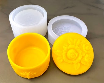 May include: Two white silicone molds for making candles or soap, one with a floral design and one plain. A yellow candle or soap with a floral design is sitting on top of the floral mold.
