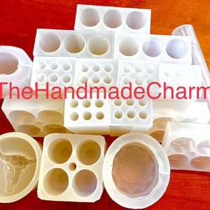 May include: A collection of white silicone molds for crafting. The molds are various shapes and sizes, including round, square, and rectangular. Some molds have multiple cavities, while others have a single cavity. The molds are arranged on a wooden surface. The text "TheHandmadeCharm" is visible in the image.