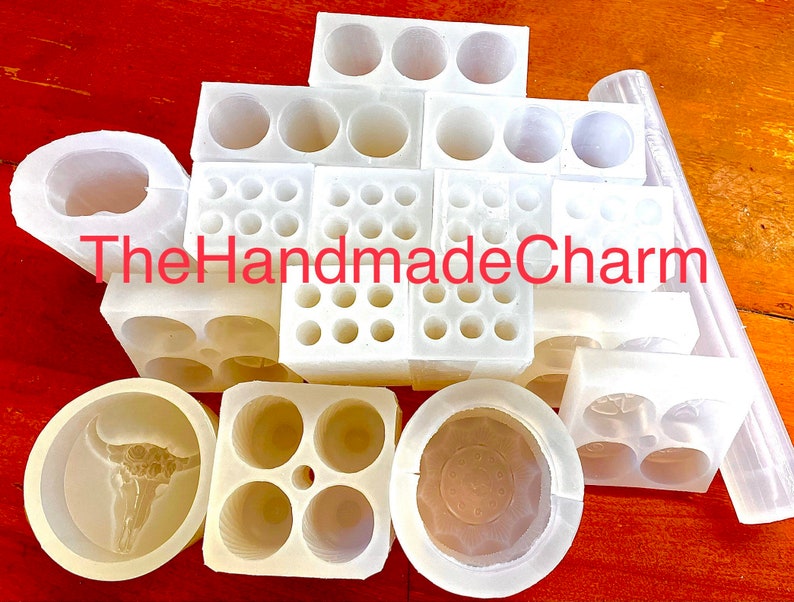 May include: A collection of white silicone molds for crafting. The molds are various shapes and sizes, including round, square, and rectangular. Some molds have multiple cavities, while others have a single cavity. The molds are arranged on a wooden surface. The text "TheHandmadeCharm" is visible in the image.