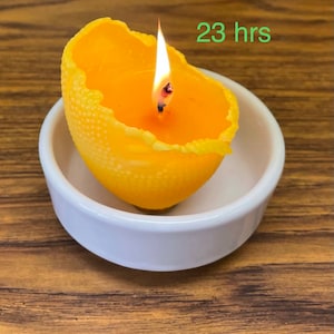 K&ouml;nnte beinhalten: Eine gelbe Zitruskerze brennt in einer wei&szlig;en Schale. Die Kerze hat eine strukturierte Oberfl&auml;che und ist mit "23 Stunden" beschriftet.