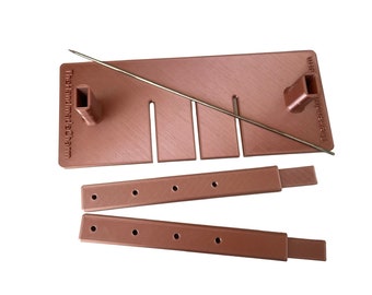 May include: A copper-coloured plastic soap cutter set featuring a rectangular base with three vertical slots, two upright rectangular posts, and a thin metal wire. Two separate rectangular bars with four circular holes each are included.