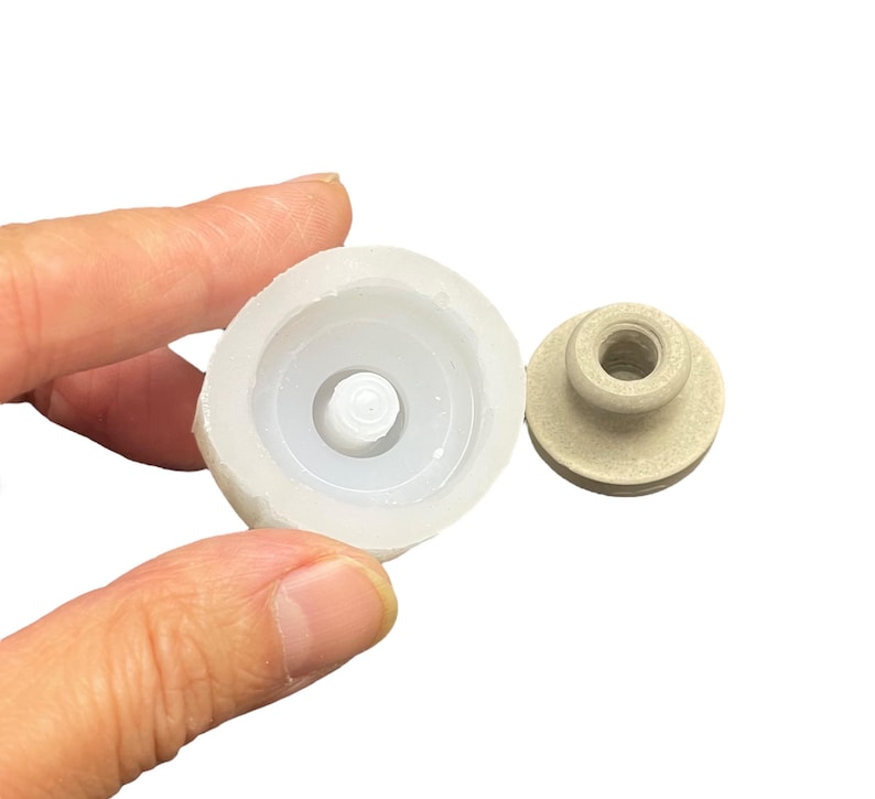 May include: A white silicone mold for making a small round object with a raised center. The mold is shown next to a beige plastic object that fits inside the mold.