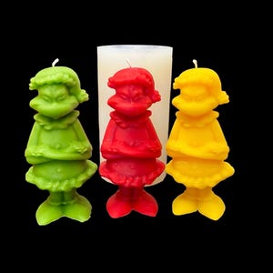 Puede incluir: Tres velas de colores con forma de personaje, verdes, rojas y amarillas. En el fondo hay un molde de vela blanco. Cada vela tiene una mecha y está sobre un fondo negro. Las velas representan un personaje.