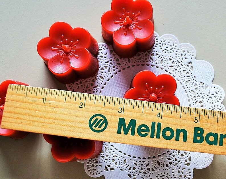 May include: Four red flower-shaped candles on a white doily with a wooden ruler that reads "Mellon Bar" across the bottom.
