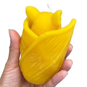 May include: A yellow beeswax candle shaped like a bat, with the bat's wings folded around its body. The candle is held in a person's hand.