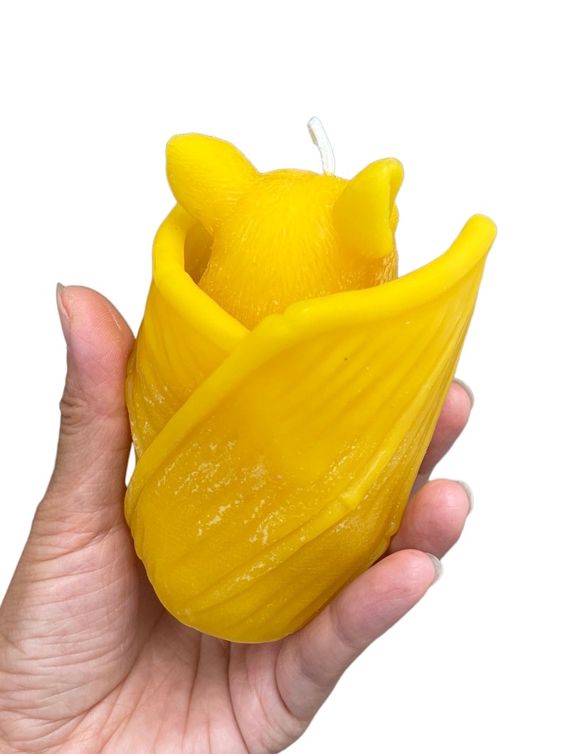 May include: A yellow beeswax candle shaped like a bat, with the bat's wings folded around its body. The candle is held in a person's hand.