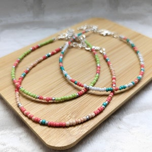 May include: Three beaded anklets in shades of pink, green, white, and teal, with silver-tone clasps. The anklets are displayed on a light brown wooden surface. The beads are small and evenly spaced, creating a delicate and colourful accessory.