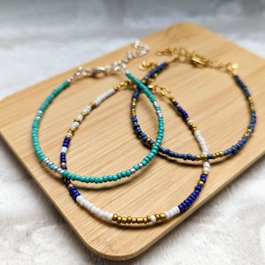May include: Three beaded bracelets with gold, white, teal, blue, and silver beads. The bracelets are on a wooden surface.