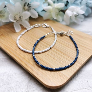 May include: Two beaded bracelets, one white and one navy blue, with silver clasps and accents. They are displayed on a light brown wooden surface, with white flowers in the background. The bracelets have a delicate, minimalist design.